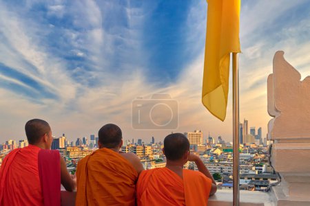 Photo for Three male Buddhist monks stand by white parapet wall of Wat Saket or Golden Mount temple and look at skyline of Bangkok, Thailand, at sunset. Orange robe, unrecognizable male adults, yellow flag, colorful cityscape, blue sky with clouds. - Royalty Free Image