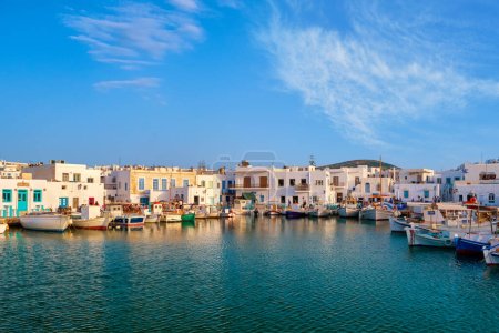 Traditional Greek fishing village and harbour on sunny day, boats moored by jetty and whitewashed houses alongside. Naoussa, Paros island, Greece. Clear blue sky, clouds, summer sunshine, colorful