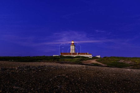 Téléchargez les photos : Vue panoramique du phare du cap Espichel, Portugal, pendant les heures de coucher du soleil. Ciel clair, peu de nuages, phare envoyant des faisceaux lumineux pour une navigation maritime sûre, peu de feuillage de terrain accidenté. - en image libre de droit