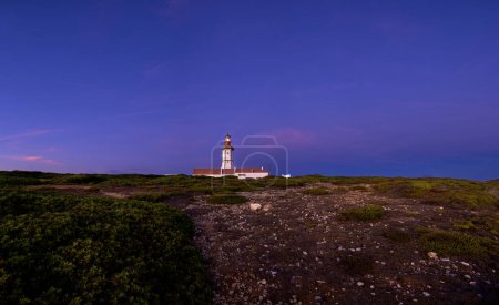Téléchargez les photos : Belle vue sur le phare du cap Espichel, Portugal, pendant les heures de coucher du soleil. Ciel clair, peu de nuages, phare envoyant des faisceaux lumineux pour une navigation maritime sûre, peu de feuillage de terrain accidenté. - en image libre de droit