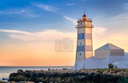 Beautiful sunset on sea shore and Santa Marta lighthouse in Cascais, Portugal. Colorful sky, clouds, sunlight, low sun, calm ocean waters, local landmark, safety and navigation tower, rocky shore. 