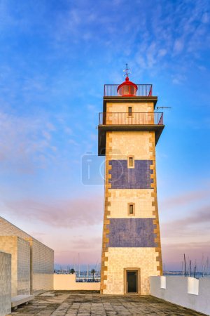 Vue du phare de Santa Marta à Cascais, Portugal au coucher du soleil. Ciel coloré, nuages, lumière du soleil, soleil bas, eaux calmes de l'océan, point de repère local, tour de sécurité et de navigation, rivage rocheux. 
