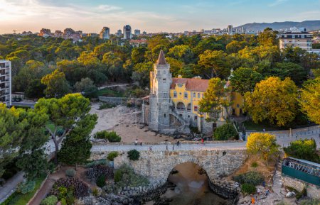 Close aerial of colorful sunset over Museum of Conde de Castro Guimaraes, Cascais, Portugal. Beautiful building in picturesque location, next to tidal pool, sandy beach, rocky shores and bridge