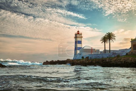 Beautiful sunset landscape on sea shore and Santa Marta lighthouse in Cascais, Portugal. Colorful sky, clouds, sunlight, low sun, calm ocean waters, local landmark, safety and navigation tower, rocky