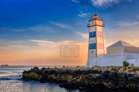Colorful sunset on sea shore and Santa Marta lighthouse in Cascais, Portugal. Colorful sky, clouds, sunlight, low sun, calm ocean waters, local landmark, safety and navigation tower, rocky shore. 