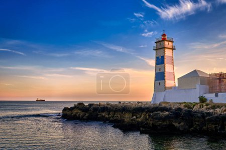 Dramatic sunset on sea shore and Santa Marta lighthouse in Cascais, Portugal. Colorful sky, clouds, sunlight, low sun, calm ocean waters, local landmark, safety and navigation tower, rocky shore. 