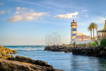 Colorful sunny day on sea shore and Santa Marta lighthouse in Cascais, Portugal. Colorful sky, clouds, sunlight, palm tree, calm ocean waters, local landmark, safety and navigation tower, rocky shore