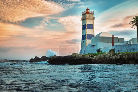 Dramatic beautiful sunset on sea shore and Santa Marta lighthouse in Cascais, Portugal. Colorful sky, clouds, sunlight, low sun, calm ocean waters, local landmark, safety and navigation tower, rocky