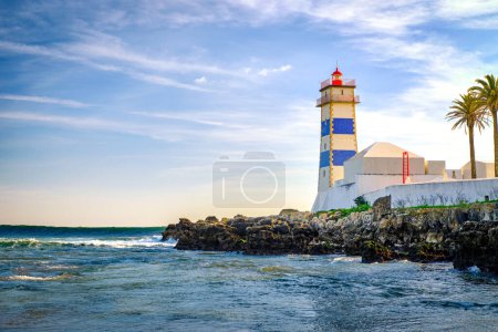 Belle soirée, sur les vagues de marée, les rivages rocheux et le phare de Santa Marta à Cascais, Portugal. Ciel bleu, nuages, lumière du soleil, soleil doux, eaux calmes de l'océan, repère local, tour de sécurité et de navigation