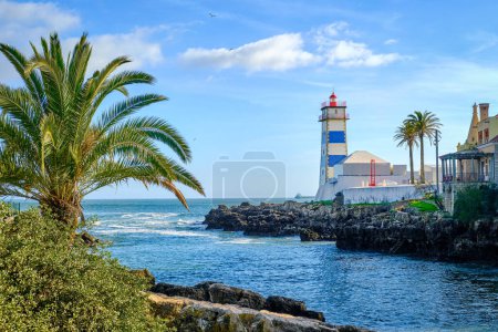 Beautiful sunny day on sea shore and Santa Marta lighthouse in Cascais, Portugal. Clear blue sky, clouds, sunlight, palm trees, calm ocean waters, local landmark, safety and navigation tower, rocky