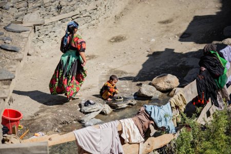 Foto de Mujer Kalash vestida con ropa tradicional haciendo la colada con su hijo cerca de su casa en la aldea Kalash, Gilgit, Pakistán - Imagen libre de derechos