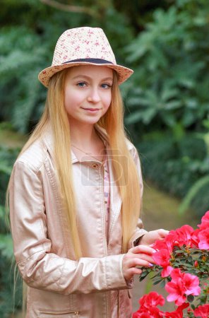 A portrait of a young blonde girl in a smiling hat stands near bushes with a pink azalea in the garden. Beauty and fashion concept