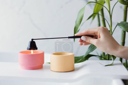 Craft pink and orange soy candles on baththube near bamboo plant at white light bathroom interior. Woman extinguish candle with black candle snuffer