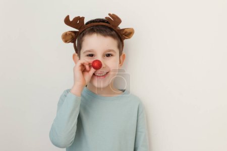Foto de Niño de escuela con cuernos de reno en la cabeza y nariz de rudolf rojo sobre un fondo blanco, espacio de copia - Imagen libre de derechos