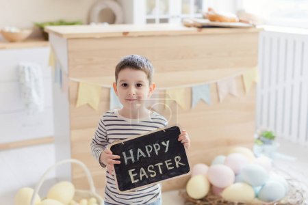 Téléchargez les photos : Garçon caucasien tient tableau avec signe de Pâques sur un intérieur de cuisine légère avec des décorations de Pâques - en image libre de droit