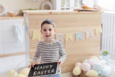 Téléchargez les photos : Garçon caucasien tient tableau avec signe de Pâques sur un intérieur de cuisine légère avec des décorations de Pâques - en image libre de droit
