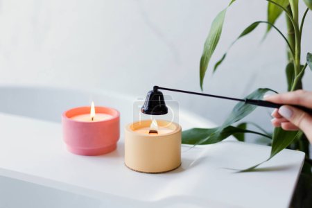 Craft pink and orange soy candles on baththube near bamboo plant at white light bathroom interior. Woman extinguish candle with black candle snuffer
