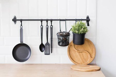 Hanging black kitchen utensils and green plant in flowerpot on a white wall background