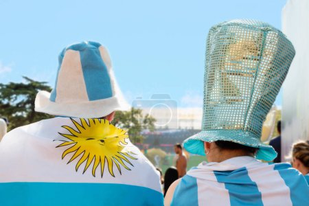 Téléchargez les photos : Fans de football hommes et femmes dos à la caméra vêtus d'un t-shirt, d'une casquette et d'un drapeau national argentin, regardez le match de l'équipe nationale argentine qui est projeté sur un écran géant dans un parc extérieur de la ville de Buenos Aires - en image libre de droit