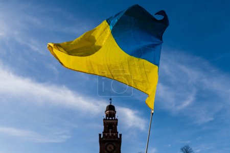 Téléchargez les photos : Drapeau aux couleurs jaunes et bleues rayées de l'Ukraine agitant dans le vent avec un ciel bleu et le soleil. En arrière-plan le Castello Sforzesco à Milan. - en image libre de droit