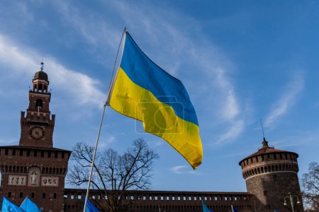 Foto de Bandera con colores a rayas amarillas y azules de Ucrania ondeando en el viento con un cielo azul y sol. En el fondo el Castello Sforzesco en Milán. - Imagen libre de derechos