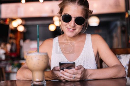 Téléchargez les photos : Portrait d'une femme heureuse utilisant un téléphone portable dans un café tout en buvant une frappe - en image libre de droit