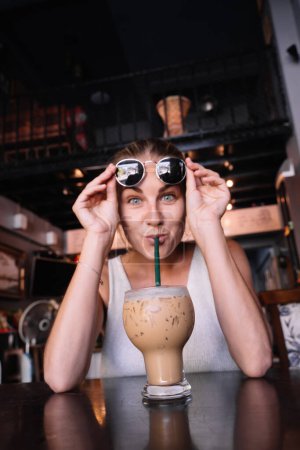 Foto de Foto vertical de una mujer divertida tomando un café helado con paja - Imagen libre de derechos