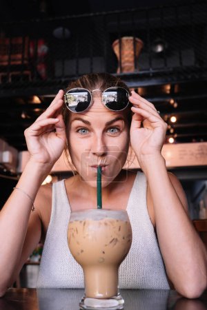 Téléchargez les photos : Photo verticale d'une femme drôle tenant des lunettes de soleil tout en buvant un café glacé avec de la paille - en image libre de droit