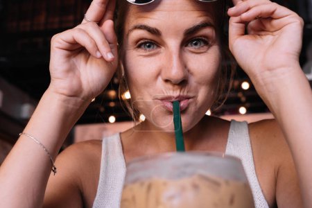 Téléchargez les photos : Gros plan portrait d'une femme blonde drôle buvant du café glacé avec une paille - en image libre de droit