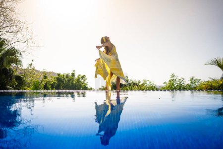 Foto de Mujer con vestido amarillo bailando junto a una piscina tropical al aire libre con la imagen reflejada en el agua - Imagen libre de derechos