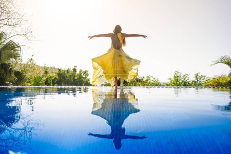 Photo for Woman with yellow dress raising arms in an outdoor pool with the image reflected on the water - Royalty Free Image