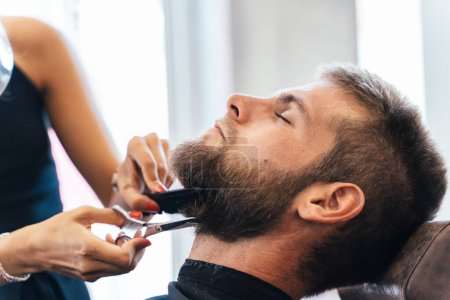 Téléchargez les photos : Coiffeur méconnaissable coupant la barbe d'un homme dans un salon - en image libre de droit