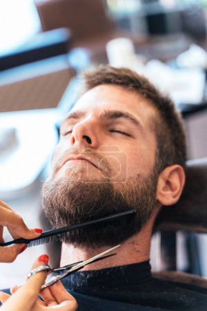 Foto de Foto vertical de cerca de un hombre relajado en una silla de un salón de belleza mientras un peluquero recorta su barba - Imagen libre de derechos