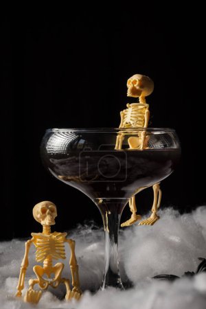 Téléchargez les photos : Gros plan de verre de veuve noire Halloween cocktail avec squelette sur toile d'araignée, fond noir, vertical - en image libre de droit