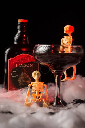 Téléchargez les photos : Gros plan de verre de cocktail de veuve noire Halloween avec squelettes et bouteille de poison sur toile d'araignée, fond noir, éclairage rouge, vertical - en image libre de droit