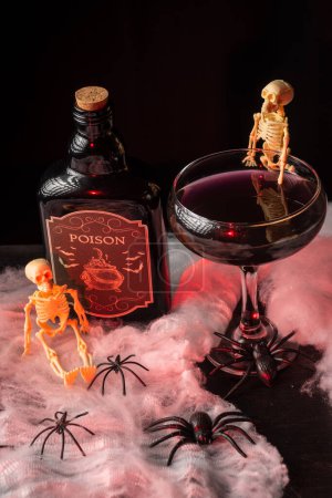 Téléchargez les photos : Vue aérienne du verre de cocktail de veuve noire Halloween avec squelettes et bouteille de poison sur toile d'araignée, illuminé en rouge, fond noir, vertical - en image libre de droit