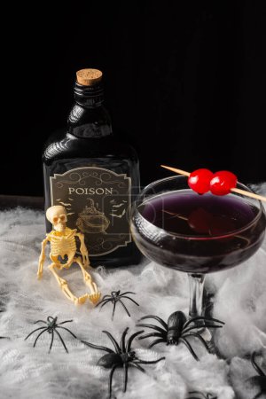 Téléchargez les photos : Vue aérienne du verre à cocktail noir Halloween avec des cerises rouges, des squelettes et une bouteille de poison sur toile d'araignée, fond noir, vertical - en image libre de droit