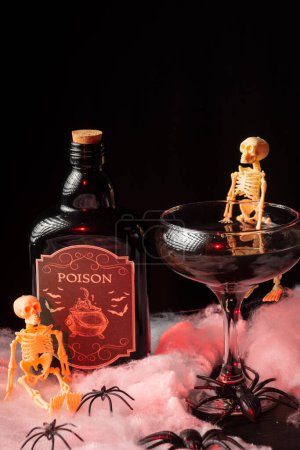 Téléchargez les photos : Vue du dessus du verre du cocktail de veuve noire d'Halloween avec squelettes et bouteille de poison sur toile d'araignée, illuminé en rouge, fond noir, vertical - en image libre de droit