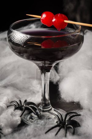 Téléchargez les photos : Gros plan de verre de cocktail de veuve noire Halloween avec des cerises rouges et des araignées sur toile d'araignée, fond noir, vertical - en image libre de droit