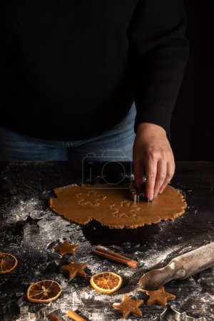 Photo for Top view of woman's hand with mold on gingerbread cookie dough on dark wooden table with cinnamon, flour, oranges and rolling pin, vertical - Royalty Free Image