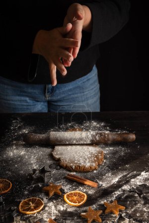 Photo for Top view of woman's hands with flour on dark table with gingerbread cookie dough with rolling pin, flour and molds, black background, vertical, with copy space - Royalty Free Image