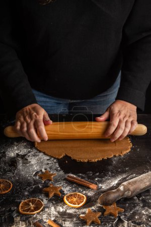 Photo for View of woman's hands with rolling pin on dark table with gingerbread cookie dough, flour, cinnamon and molds, black background, vertical - Royalty Free Image