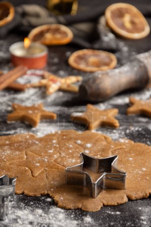 Photo for Top view of gingerbread cookie dough for Christmas with star molds, cinnamon, oranges and candle on dark table with flour, vertical - Royalty Free Image