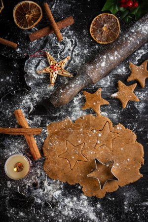 Photo for Aerial view of gingerbread cookie dough for Christmas with star molds, cinnamon and candle on dark table with flour, vertical - Royalty Free Image