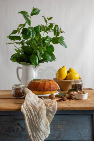 View of rustic kitchen table with bundt of lemon, walnuts, hazelnuts, cloth and jug with green bouquet, white background, vertical, with copy space