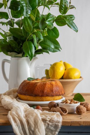 Close-up of rustic kitchen table with lemon bundt, walnuts, hazelnuts, lemons, cloth and jug with green bouquet, white background, vertical, with copy space
