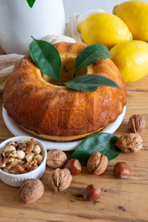 Aerial view of table with lemon bundt, walnuts, hazelnuts, lemons and cloth, white background, vertical, with copy space