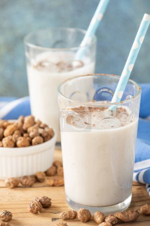 Top view of two glasses with horchata and blue straws on wooden table with tiger nuts and napkin, gray background, vertical, with copy space