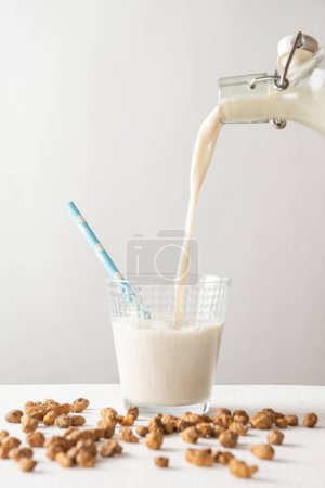 Closeup of bottle pouring horchata into glass with blue straw on white table with tiger nuts, white background, vertical, with copy space