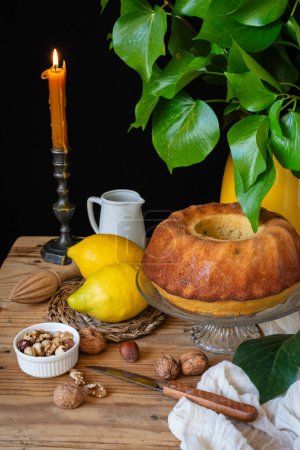 Top view of lemon bundt on wooden table with lemons, dried fruits, lit candle and vase with green leaves, black background, vertical